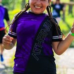 A cheerful female runner flashes a peace sign during an outdoor run with others in a sunny, green setting.