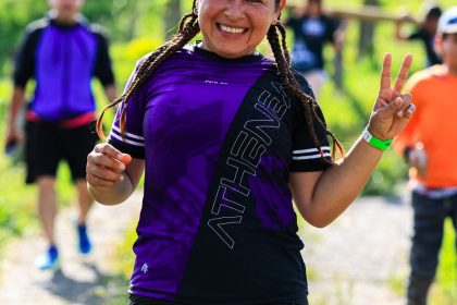 A cheerful female runner flashes a peace sign during an outdoor run with others in a sunny, green setting.