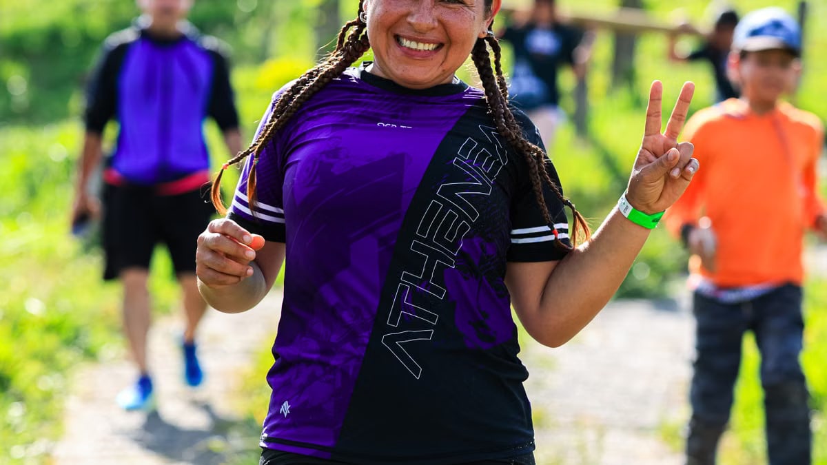 A cheerful female runner flashes a peace sign during an outdoor run with others in a sunny, green setting.